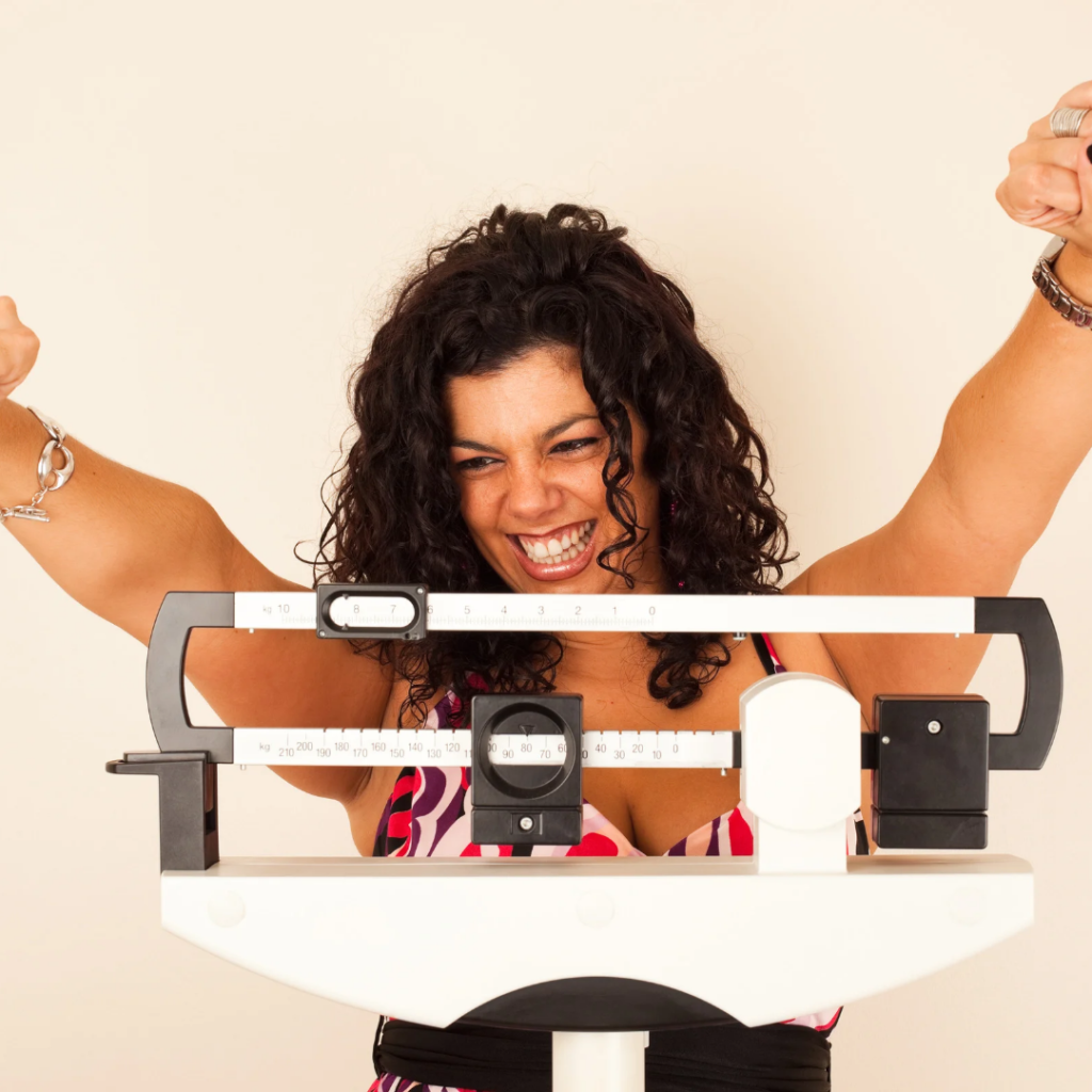 A smiling woman celebrating her sustainable weight loss progress as she steps onto a balance beam scale.
