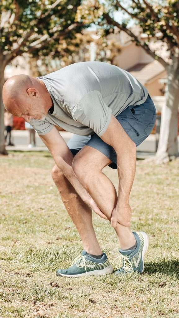 An athletic runner over 50 kneeling to gently check and massage a sore calf and ankle outdoors, illustrating how to listen to early warning signs and joint discomfort.
