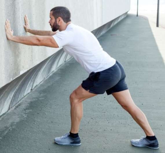 A side view of a man standing facing a wall, leaning forward to perform a calf stretch for routine foot care, with his back leg straight and heel pressed firmly into the floor.