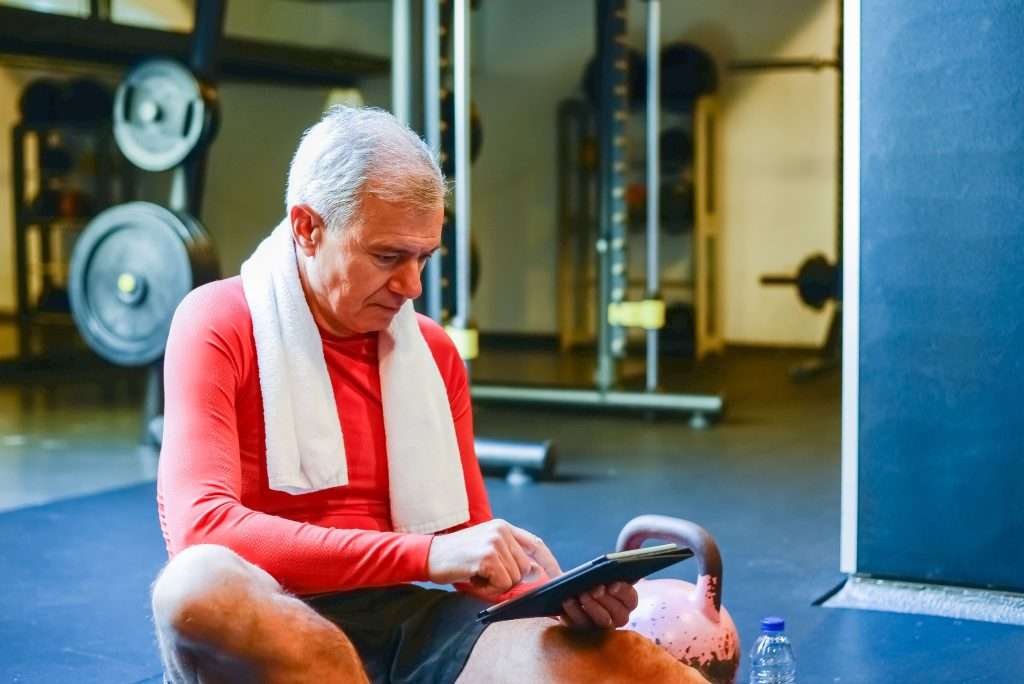 An older man in a gym sitting on a mat and looking at a tablet to guide his consistent fitness routine.