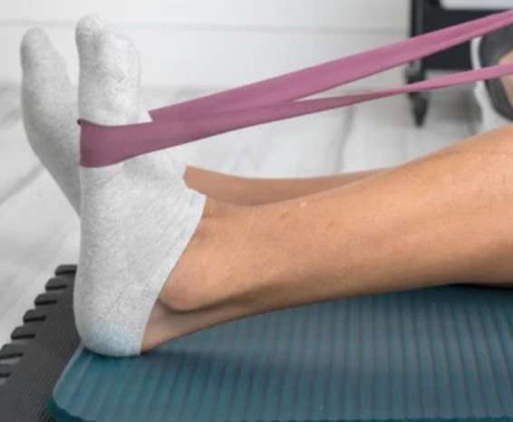 A close-up of a person sitting on the floor performing a towel stretch for foot flexibility, looping a resistance band around the ball of the foot and gently pulling back to stretch the arch and calf.